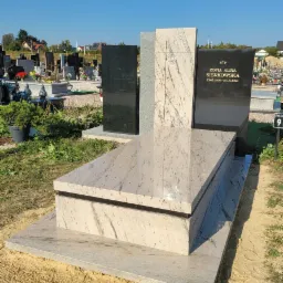 Nowoczesny, jasny pomnik z ciemnymi elementami na cmentarzu, z inskrypcją i wyrzeźbioną różą, na tle innych grobów i zabudowań.