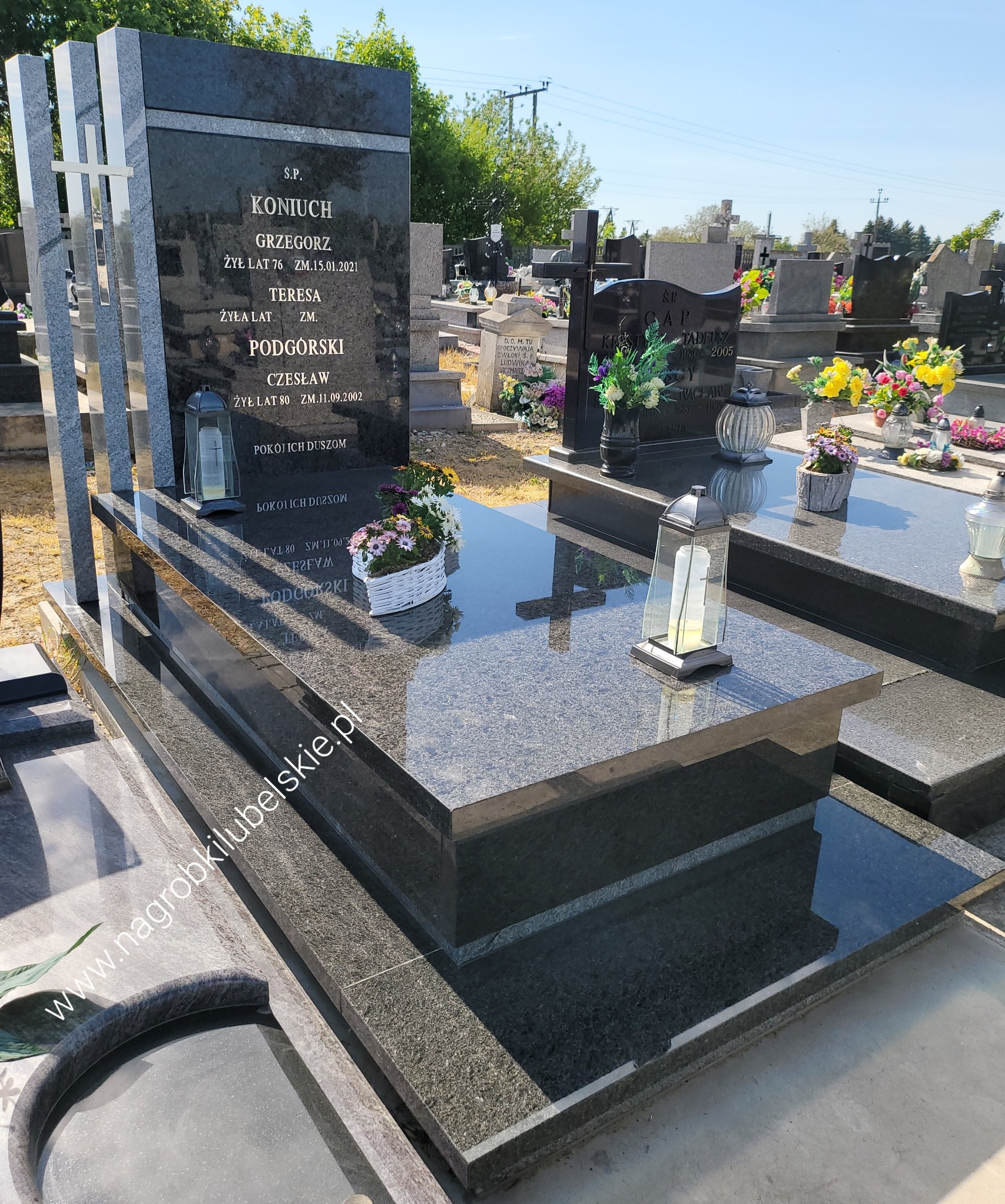 Granitowy pomnik z ciemnego kamienia z wysokimi tablicami pionowymi, na cmentarzu, z widocznymi kwiatami w białym koszyczku i lampionem ze świecą.