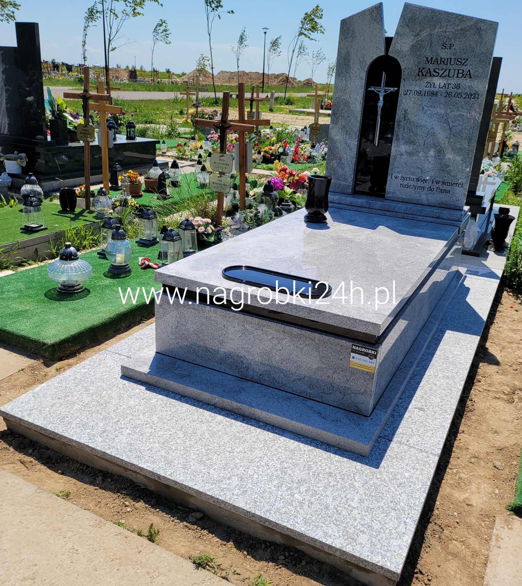 Jasnoszary, nowoczesny pomnik z krzyżem w niszy, na cmentarzu z licznymi nagrobkami i kwiatami w tle.