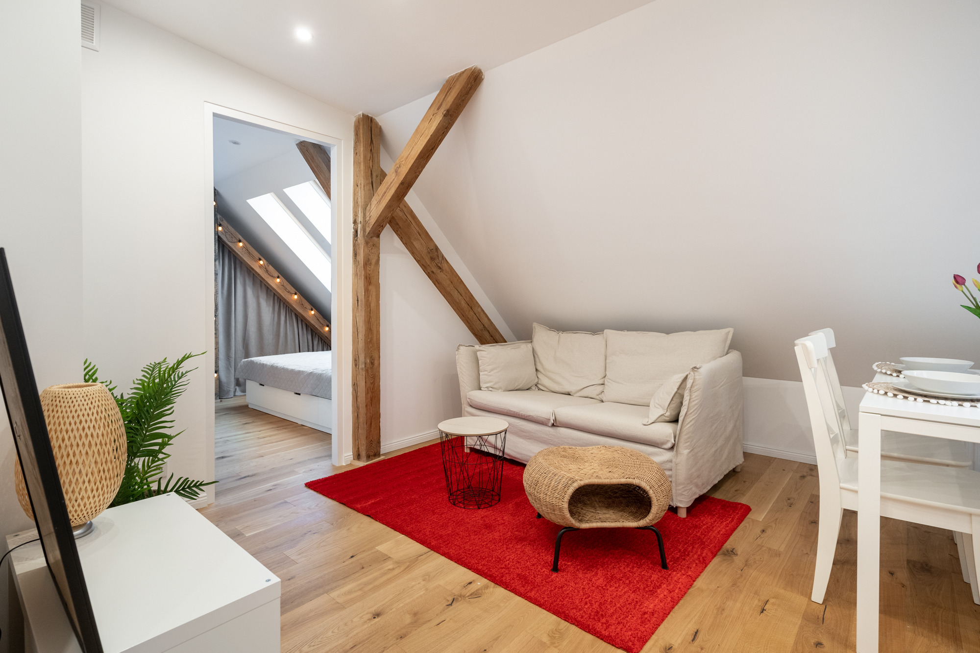 Jasne poddasze z drewnianymi belkami konstrukcyjnymi, czerwonym dywanem, beżową sofą i widokiem na sypialnię z oknami dachowymi.