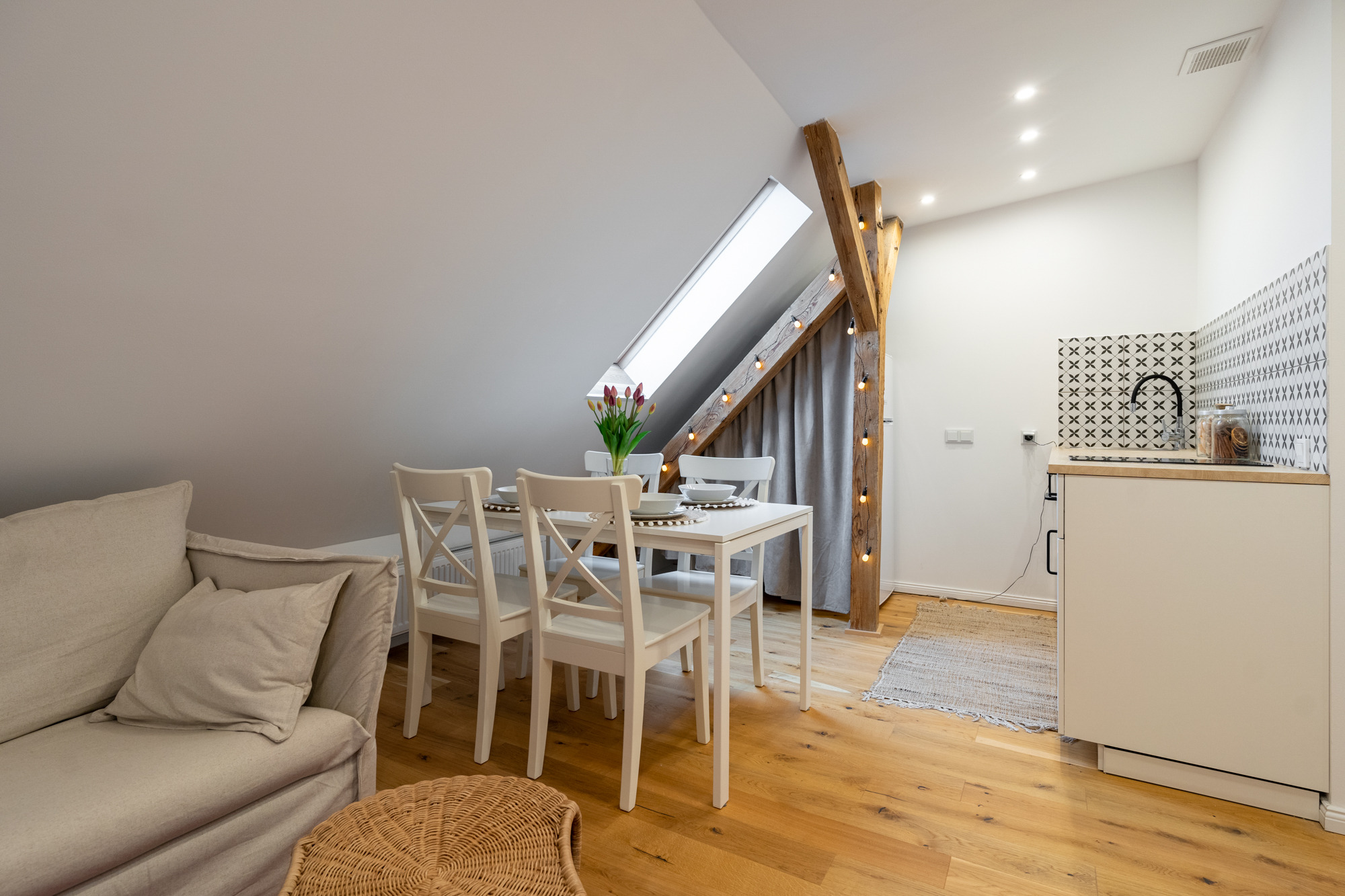 Jasne poddasze z drewnianą podłogą, sofą, stołem z kwiatami i aneksem kuchennym z wzorzystymi płytkami.