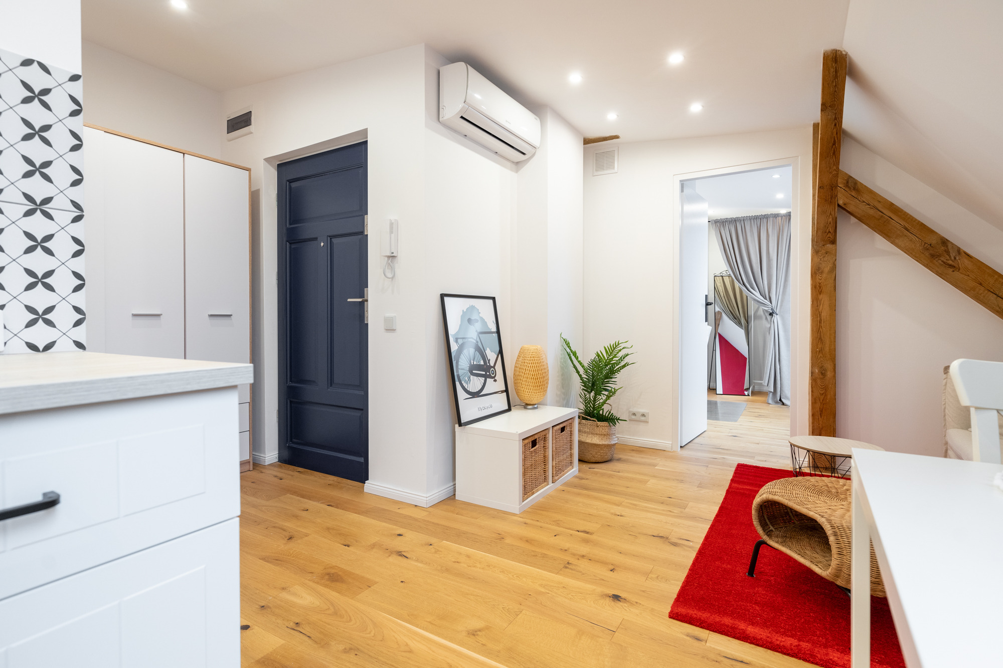 Jasne wnętrze poddasza z drewnianą podłogą, granatowymi drzwiami, białymi meblami, klimatyzatorem i dekoracjami, w tym obrazkiem roweru i rośliną w wiklinowym koszu.