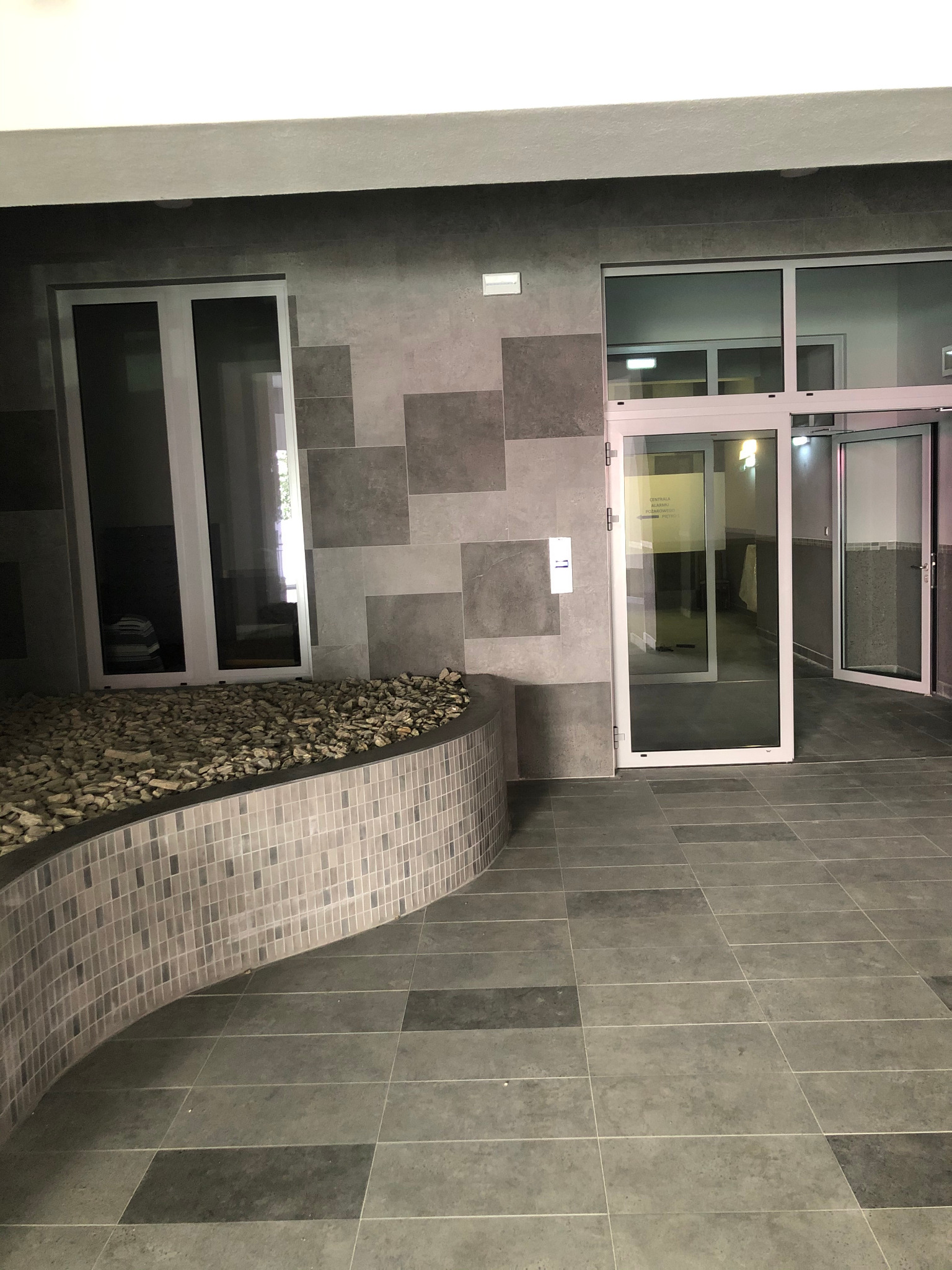 Szare, kwadratowe płytki podłogowe i ścienne ułożone w geometryczny wzór, z niskim murem z mozaiki i kamieniami dekoracyjnymi, oraz otwarte szklane drzwi wejściowe.