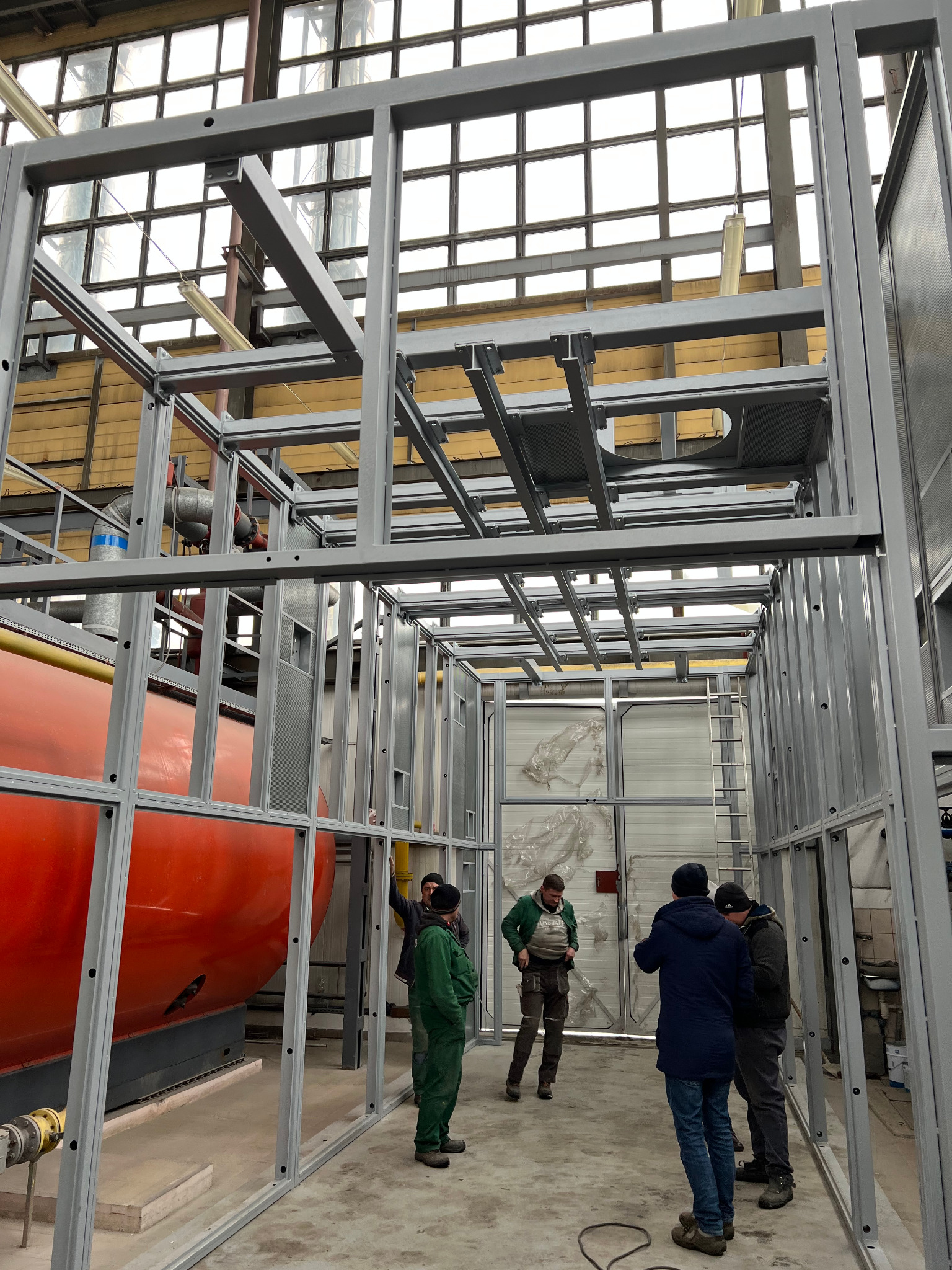 Montaż metalowej konstrukcji w hali przemysłowej, widoczny duży pomarańczowy zbiornik i grupa robotników w ubraniach roboczych.