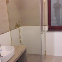 USŁUGI OGÓLNOBUDOWLANE MARCIN M - Nowoczesna kabina prysznicowa z złotymi elementami i szklanymi drzwiami, umywalka na drewnianym blacie, widok na okno z czerwoną ramą, minimalistyczny design łazienki.