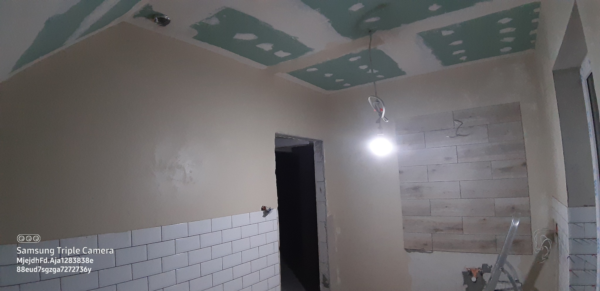 Wnętrze w trakcie prac: częściowo wykończona ściana z białych płytek, fragment ściany z imitacją drewna, sufit z widocznymi płytami kartonowo-gipsowymi i miejscami po szpachlowaniu, wisząca żarówka.