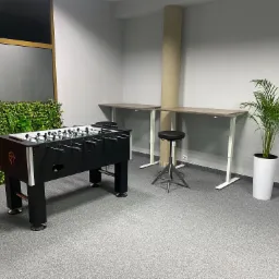 Pok&oacute;j biurowy z piłkarzykami, regulowanymi biurkami, stołkiem barowym i roślinami doniczkowymi, utrzymany w szarej kolorystyce.