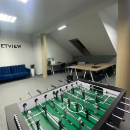 Wnętrze biura z piłkarzykami na pierwszym planie, w tle logo firmy Netview na ścianie, niebieska sofa i biurka z krzesłami.