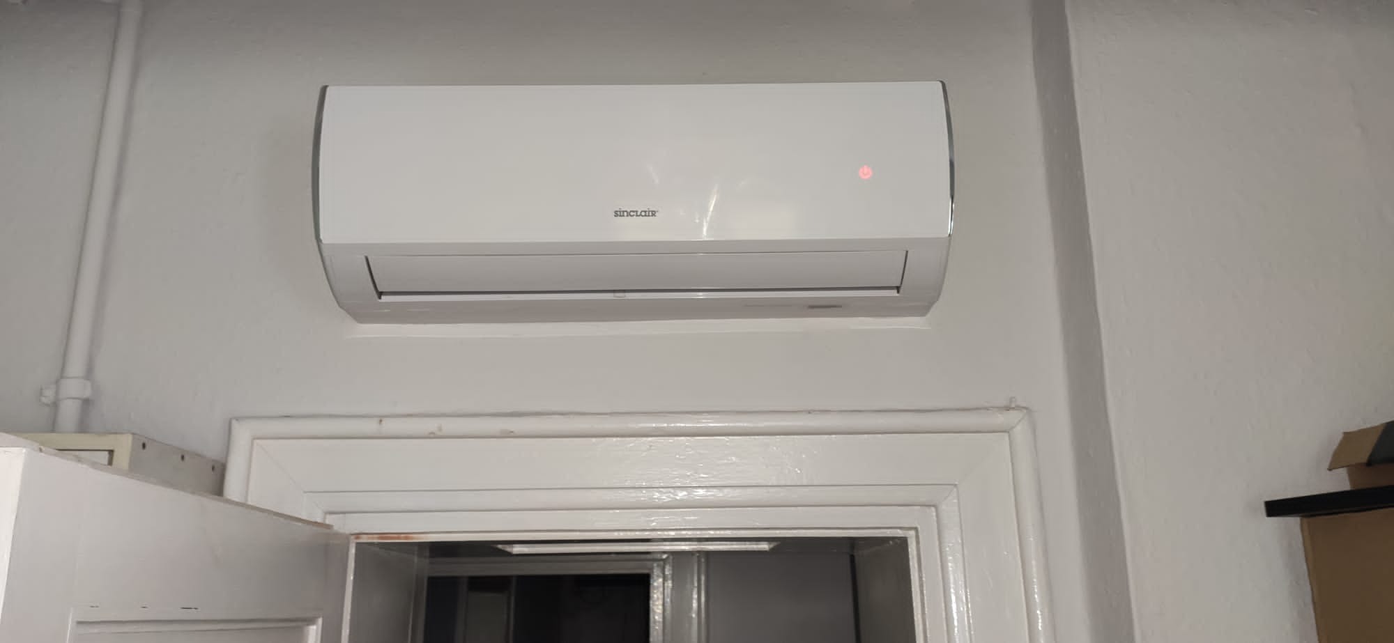 Biały klimatyzator Sinclair zamontowany na ścianie nad ościeżnicą drzwi, widoczna dioda sygnalizacyjna.