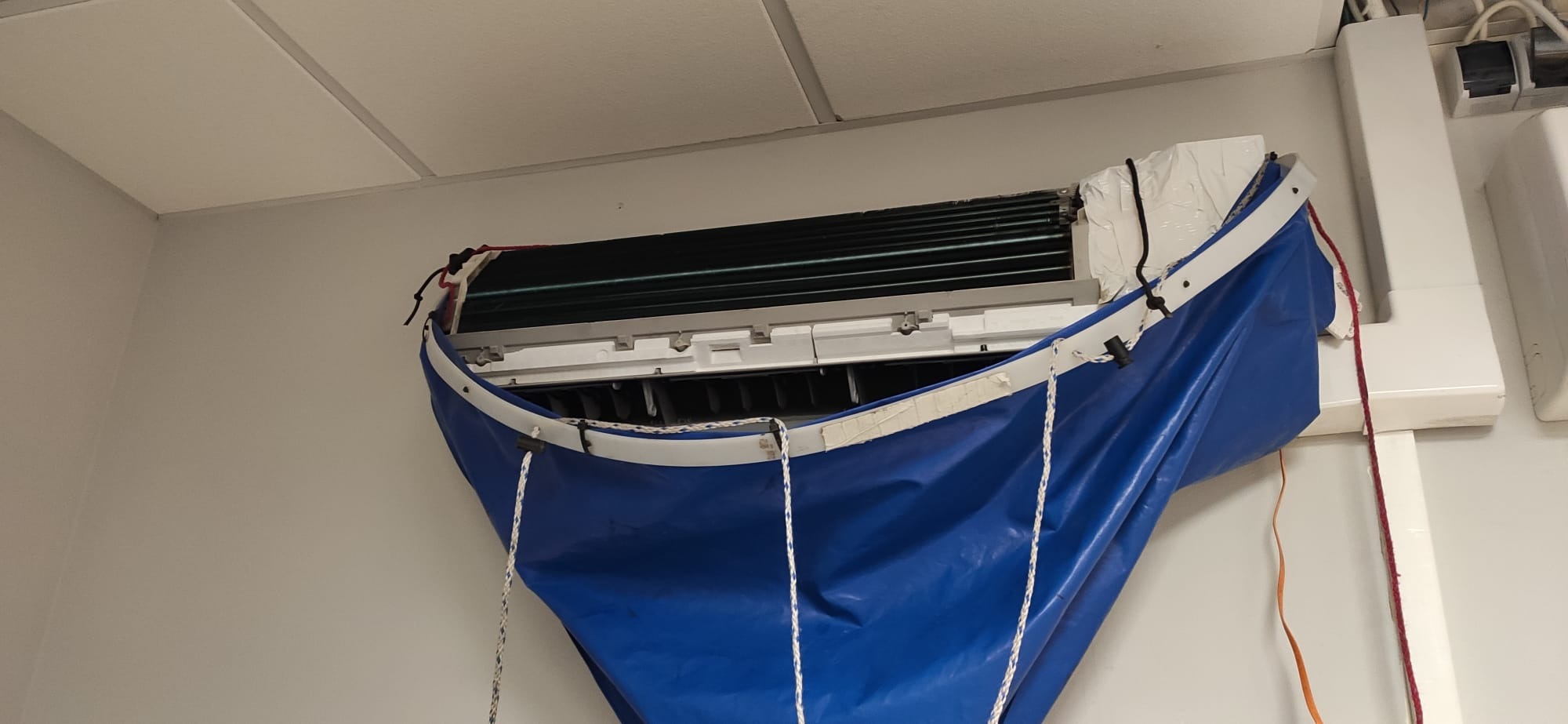 Klimatyzator ścienny zabezpieczony niebieską płachtą z białym obramowaniem i linami, prawdopodobnie w trakcie czyszczenia lub serwisu.