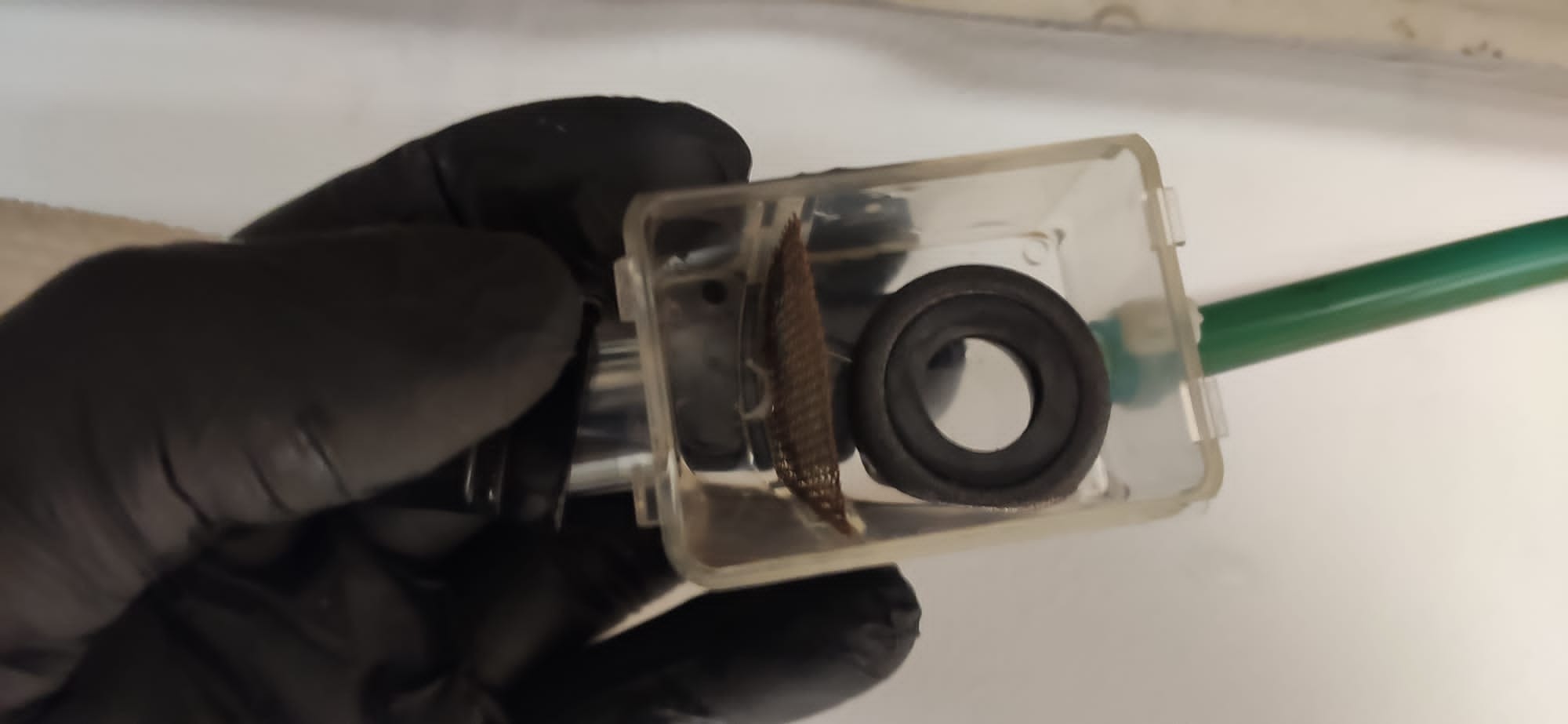 Filtr powietrza w przezroczystej obudowie, trzymany w dłoni w czarnej rękawicy, obok widoczny fragment zielonej rurki.