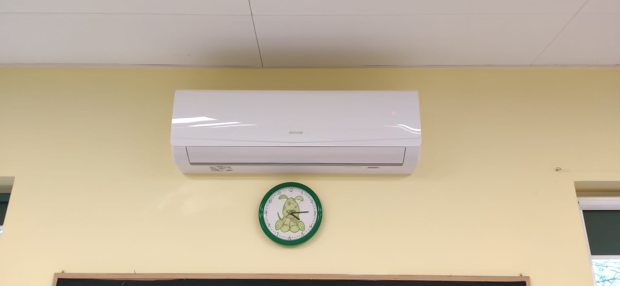 Biały klimatyzator ścienny zamontowany nad zielonym zegarem z motywem psa na jasnożółtej ścianie.