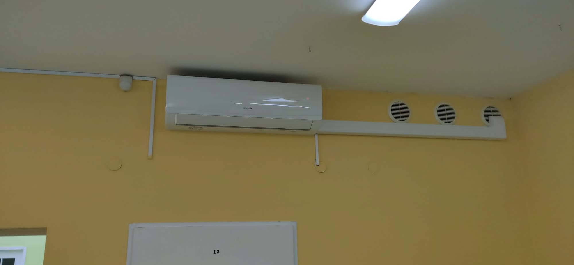 Biały klimatyzator ścienny zamontowany w biurze, widoczne okrągłe kratki wentylacyjne i żółte ściany.