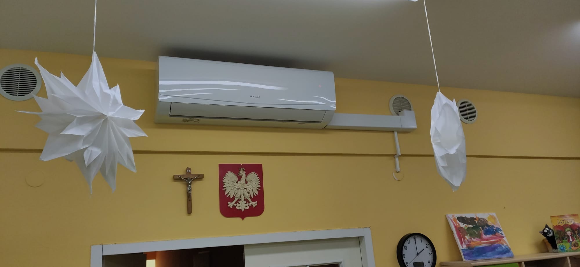 Biały klimatyzator Sinclair zamontowany na żółtej ścianie, ozdobiony papierowymi dekoracjami, krzyżem i godłem Polski.