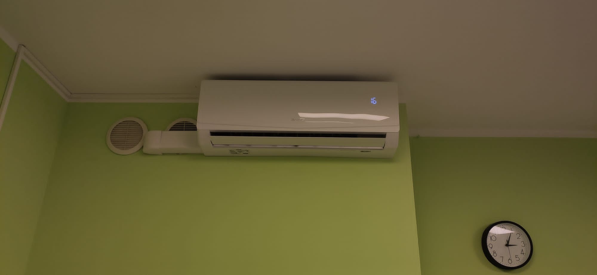 Biały klimatyzator ścienny z wyświetlaczem temperatury 16 stopni, zamontowany w pomieszczeniu o pistacjowych ścianach, obok widoczna okrągła kratka wentylacyjna i zegar ścienny.