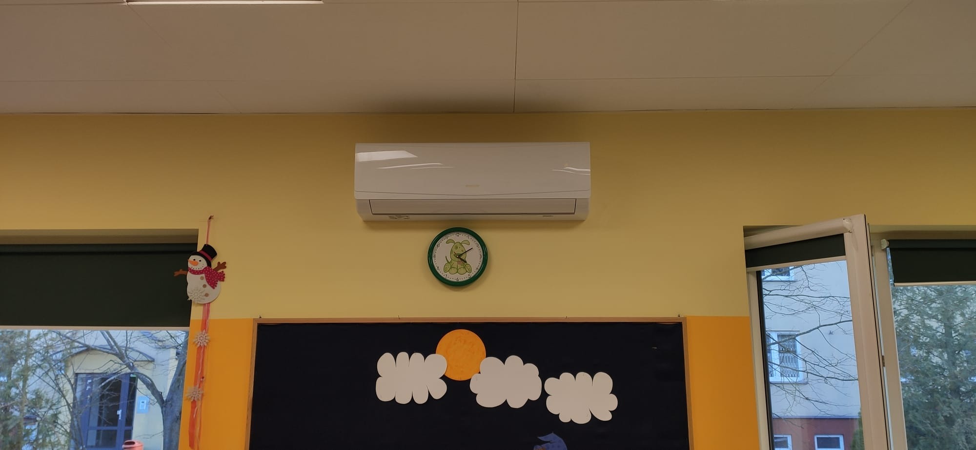Biały klimatyzator ścienny zamontowany w pokoju dziecięcym z żółtą ścianą, obok zegar z wesołym psem i tablica z rysunkami chmur i słońca.