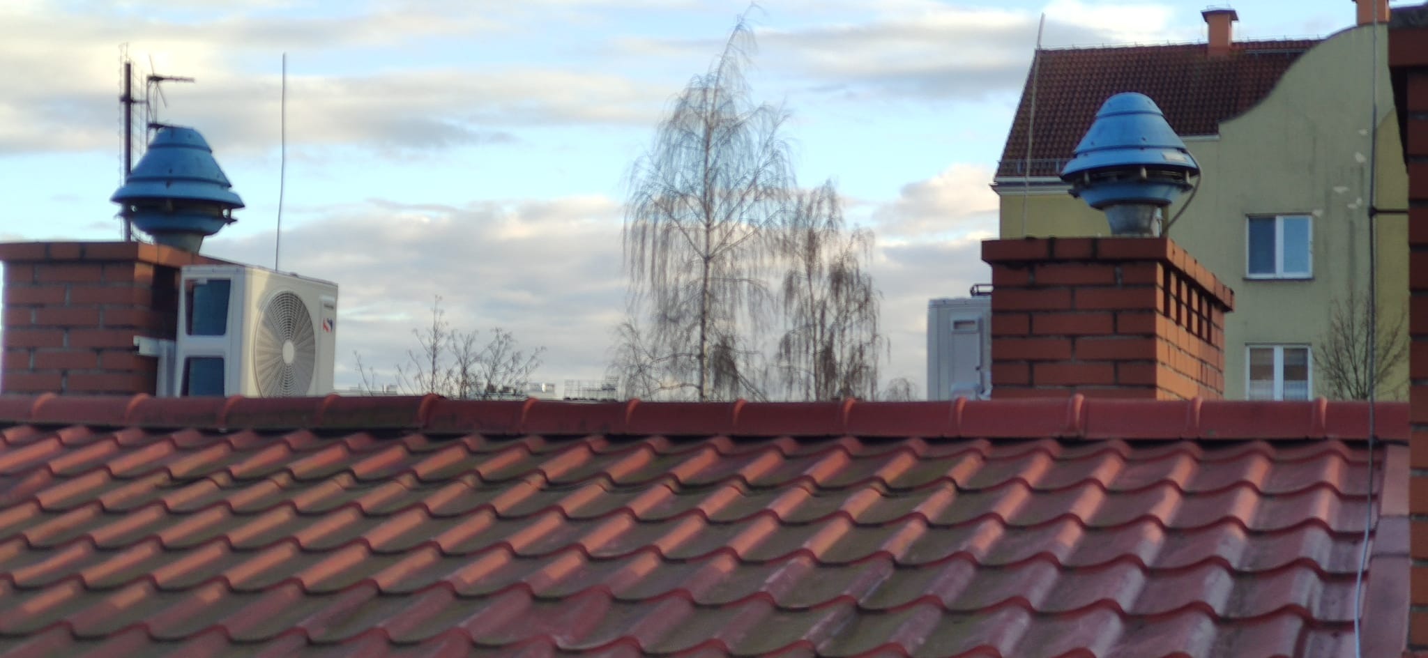 Widok z góry na dach z czerwonej dachówki, komin z niebieską nasadą wentylacyjną i jednostkę zewnętrzną klimatyzacji na tle błękitnego nieba z chmurami oraz drzewa bez liści.