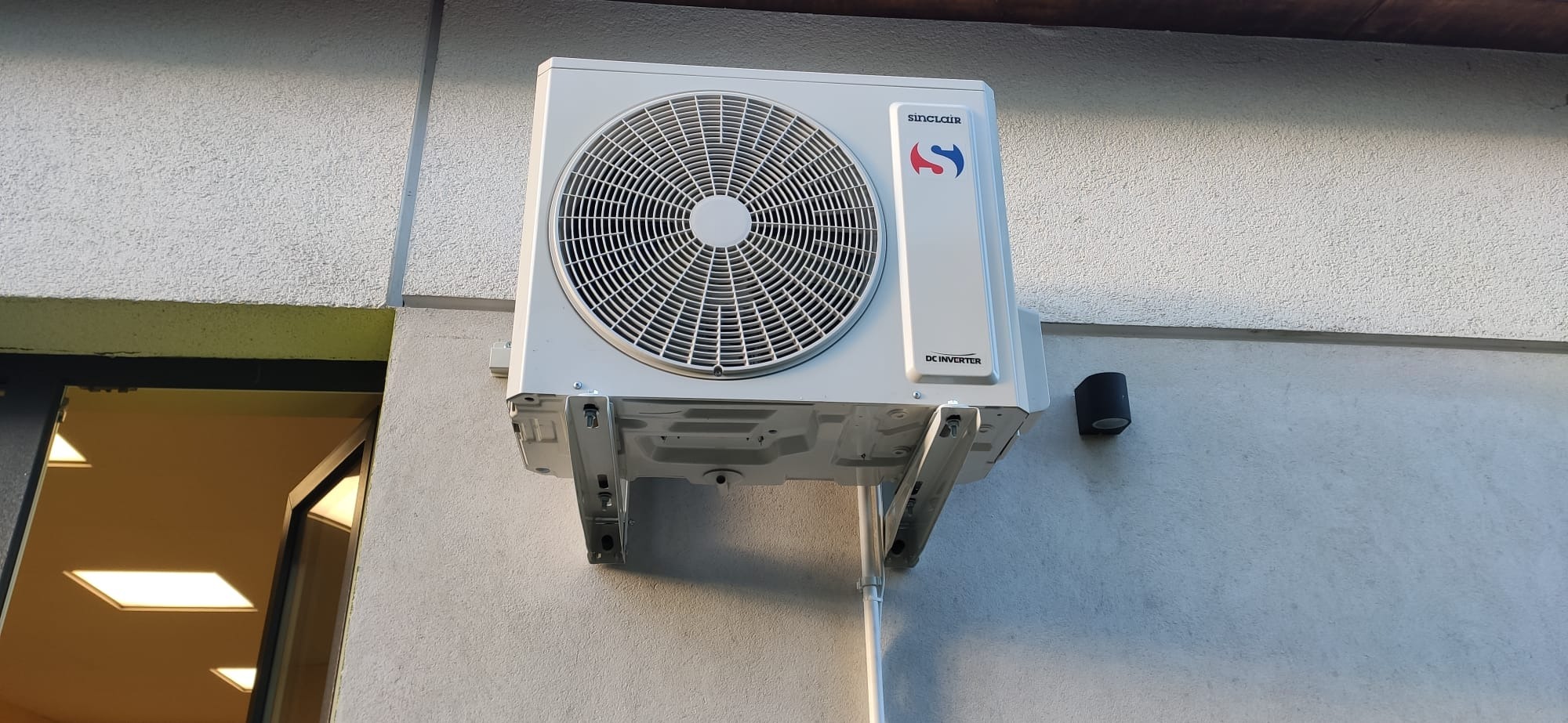 Zewnętrzna jednostka klimatyzacji Sinclair zamontowana na ścianie budynku z widoczną elewacją o fakturze baranka i czarną lampą elewacyjną obok.