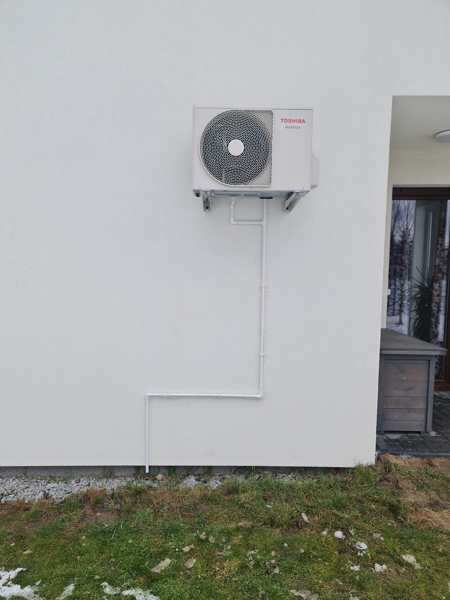 Zewnętrzna jednostka klimatyzatora Toshiba Inverter zamontowana na białej elewacji budynku z widocznymi białymi rurami instalacyjnymi.