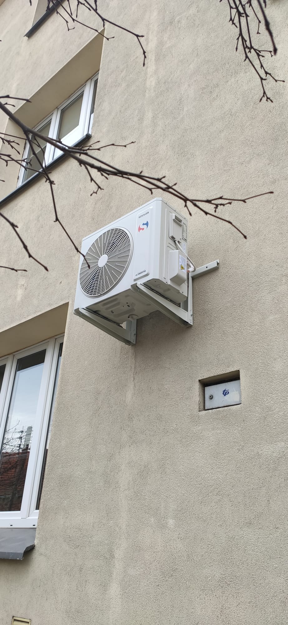Biała jednostka zewnętrzna klimatyzatora Sinclair zamontowana na elewacji budynku, widoczne gałęzie drzewa w kadrze.
