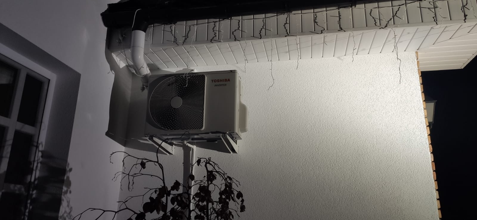 Zewnętrzna jednostka klimatyzacji Toshiba Inverter zamontowana na ścianie budynku z widoczną instalacją odprowadzającą skropliny, oświetlona girlandą światełek, częściowo zasłonięta przez gałęzie...
