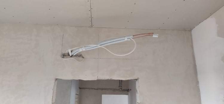 Wyprowadzone przewody i rury instalacyjne ze ściany w surowym stanie, widoczne miedziane rury i peszle na tle szarego tynku i płyty kartonowo-gipsowej na suficie.