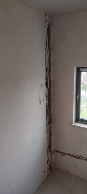 Ściana w trakcie montażu instalacji wentylacyjnej z widocznymi przewodami w wyżłobieniu, surowy tynk, okno z ciemną ramą.