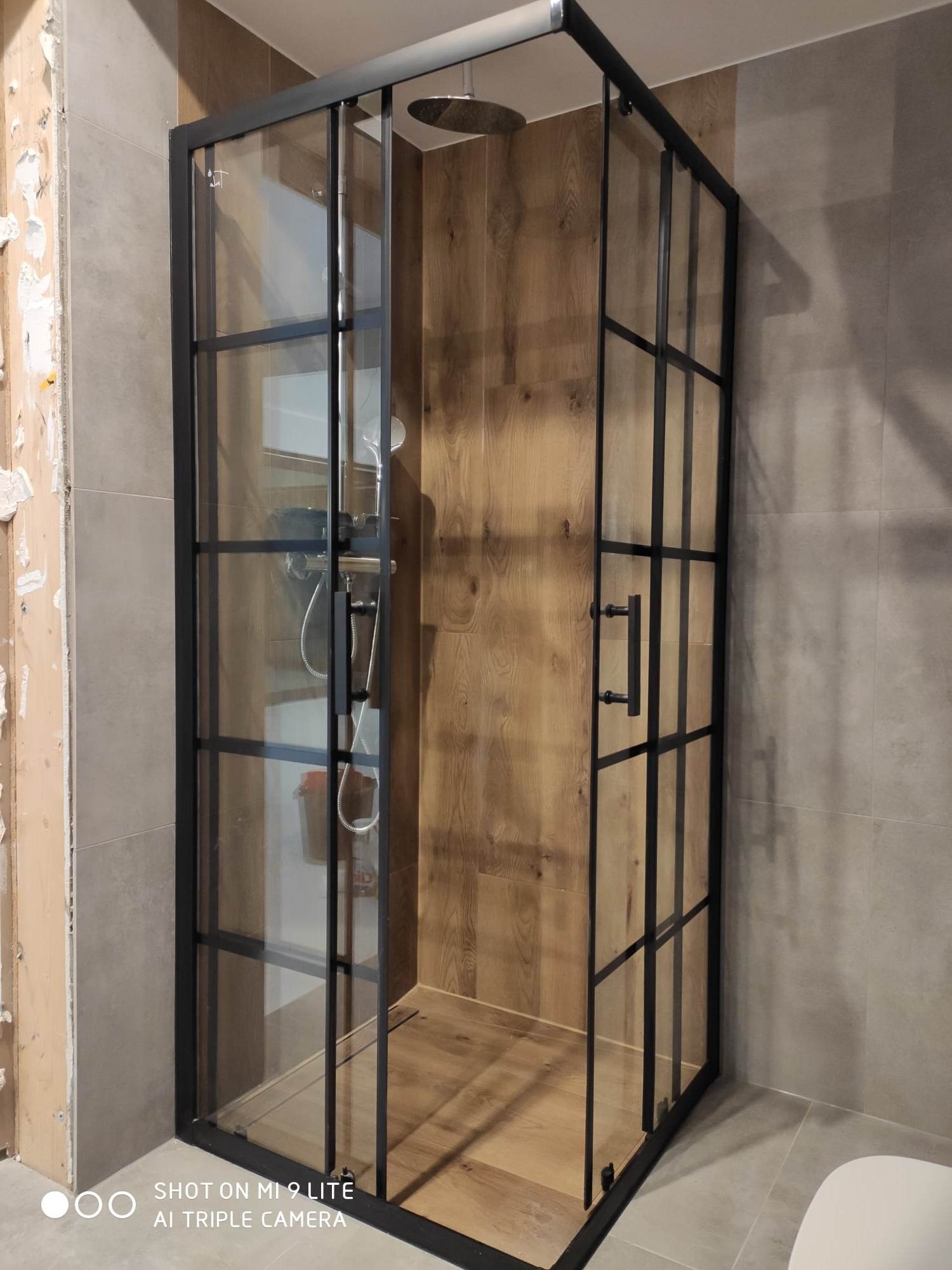 Kabina prysznicowa z czarnymi profilami i szklanymi ściankami, zamontowana w łazience z drewnopodobnymi płytkami na ścianie wewnątrz i szarymi płytkami na zewnątrz, widoczne ślady po usuniętych...
