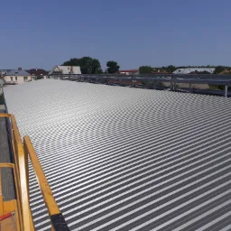 Szeroki widok na srebrne, blaszane pokrycie dachu przemysłowego z widocznymi elementami konstrukcyjnymi i częścią podnośnika koszowego po lewej stronie, na tle zabudowań mieszkalnych i błękitnego...