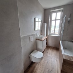 thermal komfort - Nowoczesna łazienka po remoncie w Połczynie-Zdroju. Płytki imitujące drewno na podłodze, szare płytki na ścianach, wisząca toaleta i wanna z parawanem.