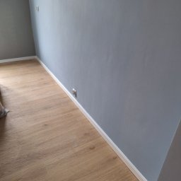 thermal komfort - Pomalowane na szaro ściany, drewniana podłoga i białe listwy przypodłogowe w pustym pokoju z gniazdkiem elektrycznym. Nowoczesne wykończenie wnętrza.