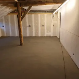 Wnętrze poddasza w trakcie prac: wylewka betonowa na podłodze, ściany pokryte płytami kartonowo-gipsowymi z widocznymi puszkami elektrycznymi, drewniany słup konstrukcyjny, surowy strop...