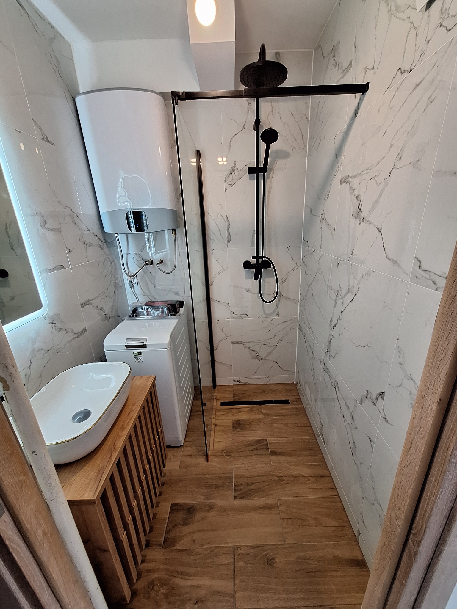 Nowoczesna łazienka z marmurowymi płytkami, kabiną prysznicową typu walk-in z czarną armaturą, umywalką na drewnianej szafce i bojlerem nad pralką.