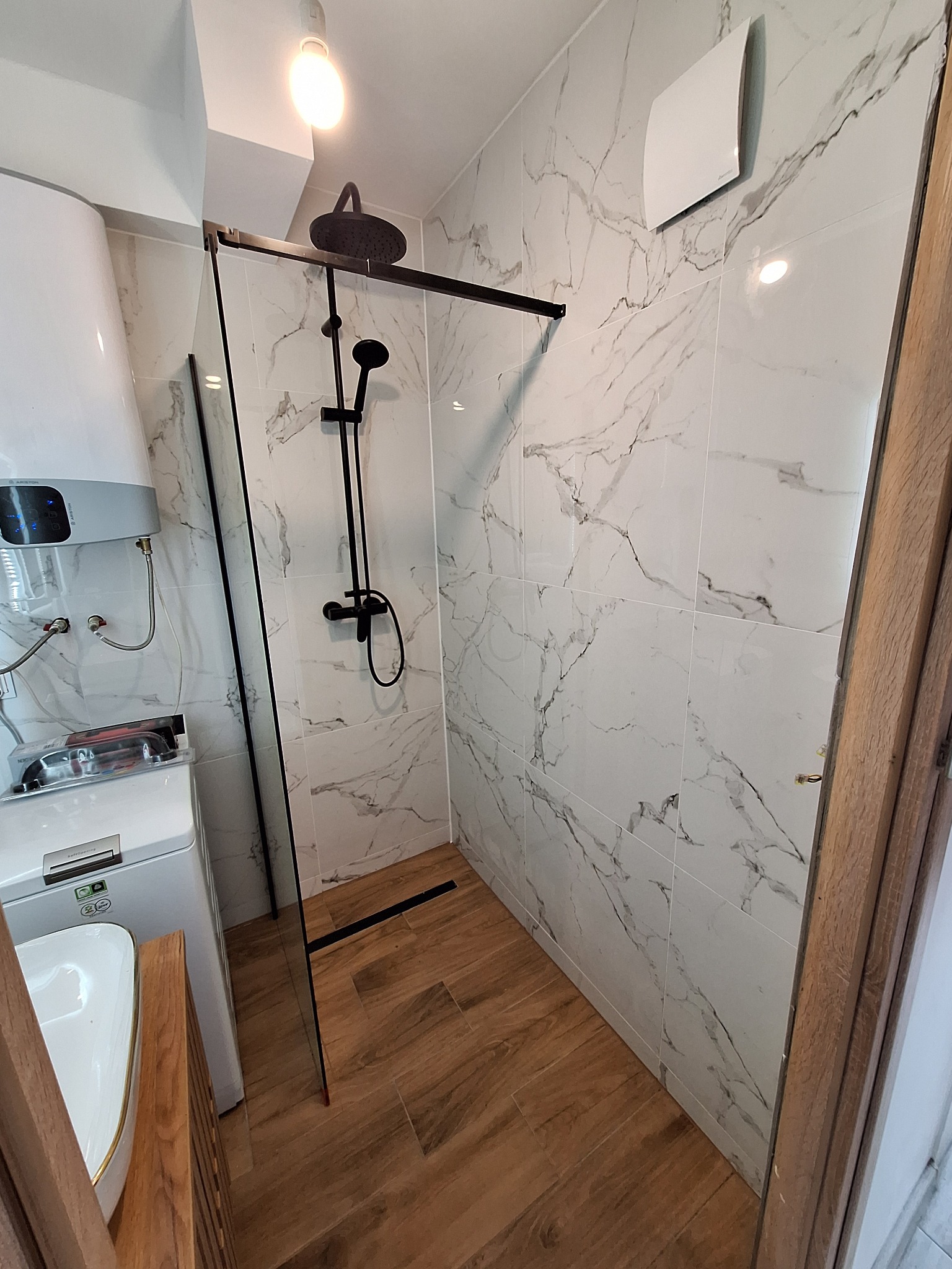 Wykończona łazienka z kabiną typu walk-in, armaturą w czarnym macie i płytkami imitującymi marmur. Widoczny bojler i pralka w zabudowie. Oświetlenie sufitowe.