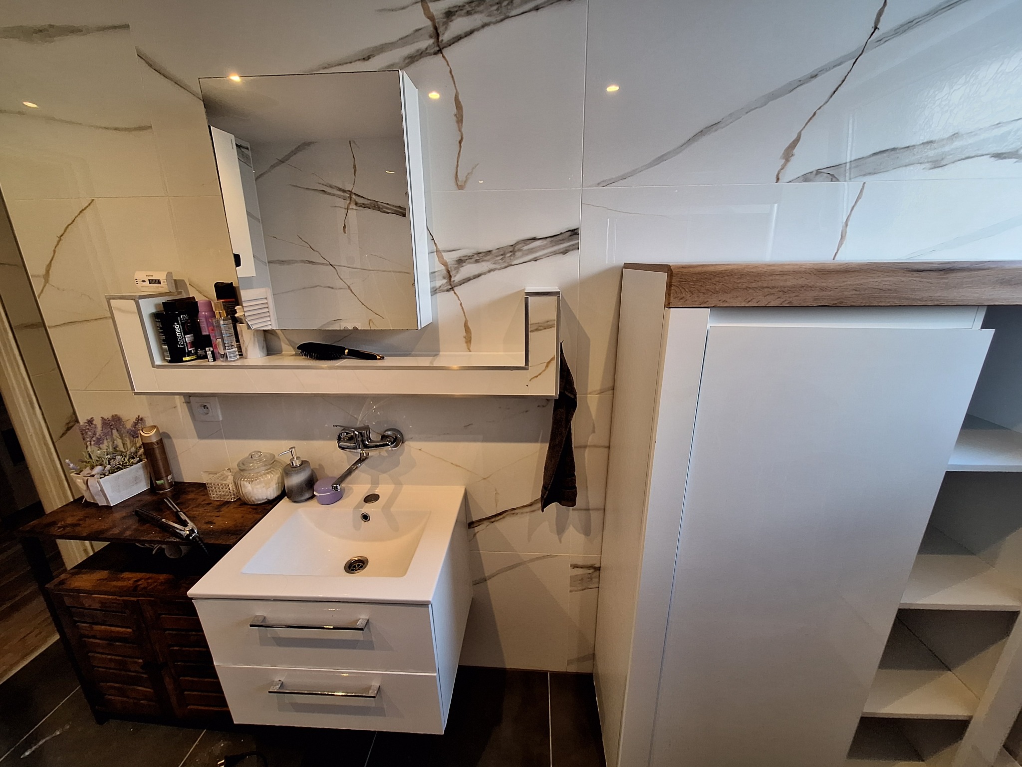 Nowoczesna łazienka z białą umywalką, szafką z drewnianym blatem i lustrem. Ściany wyłożone białymi płytkami z marmurkowym wzorem. Elementy dekoracyjne dodają przytulności.