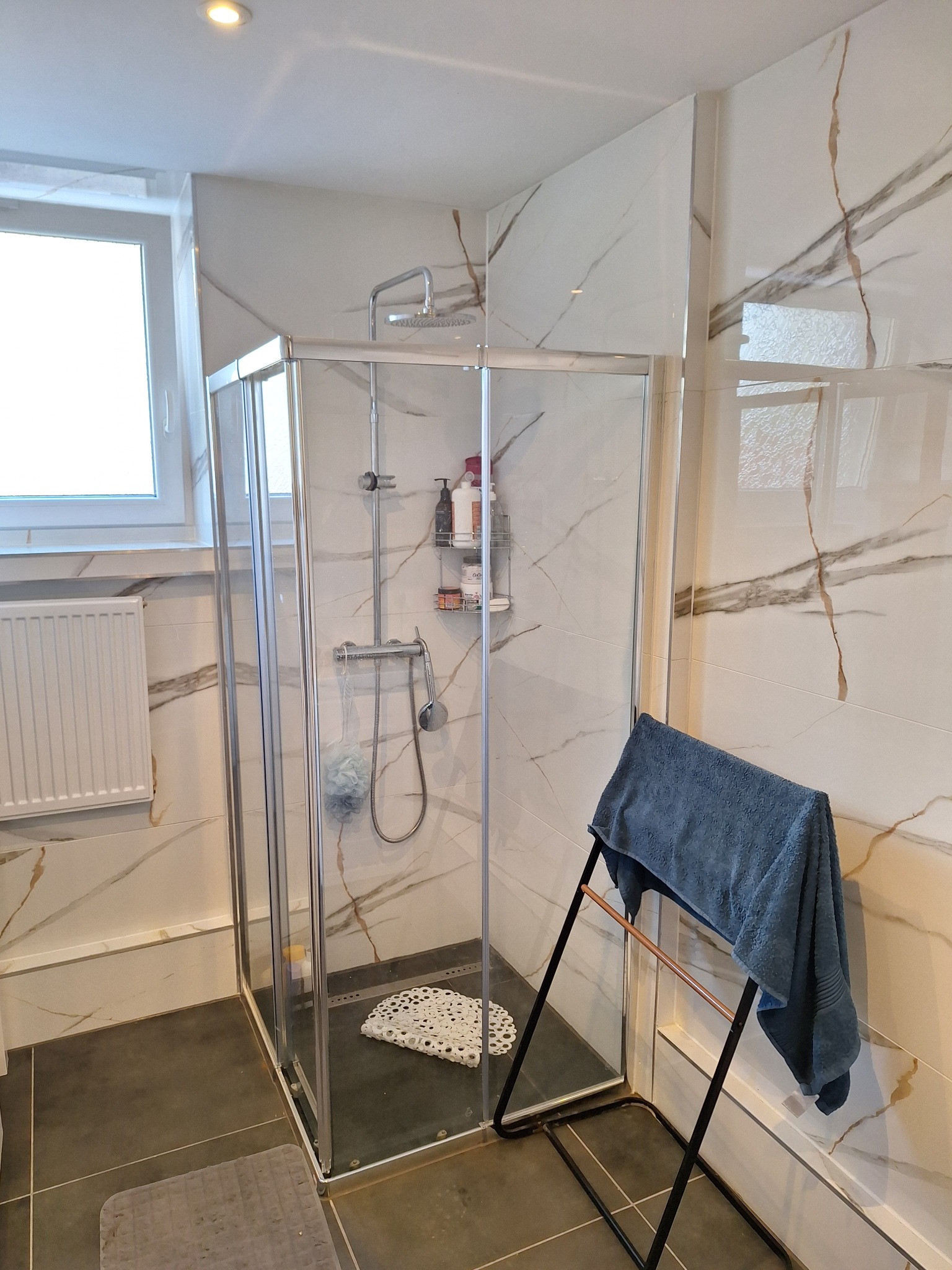 Nowoczesna kabina prysznicowa z deszczownicą, wykończona marmurowymi płytkami z brązowymi żyłami. Na zewnątrz wieszak na ręczniki.