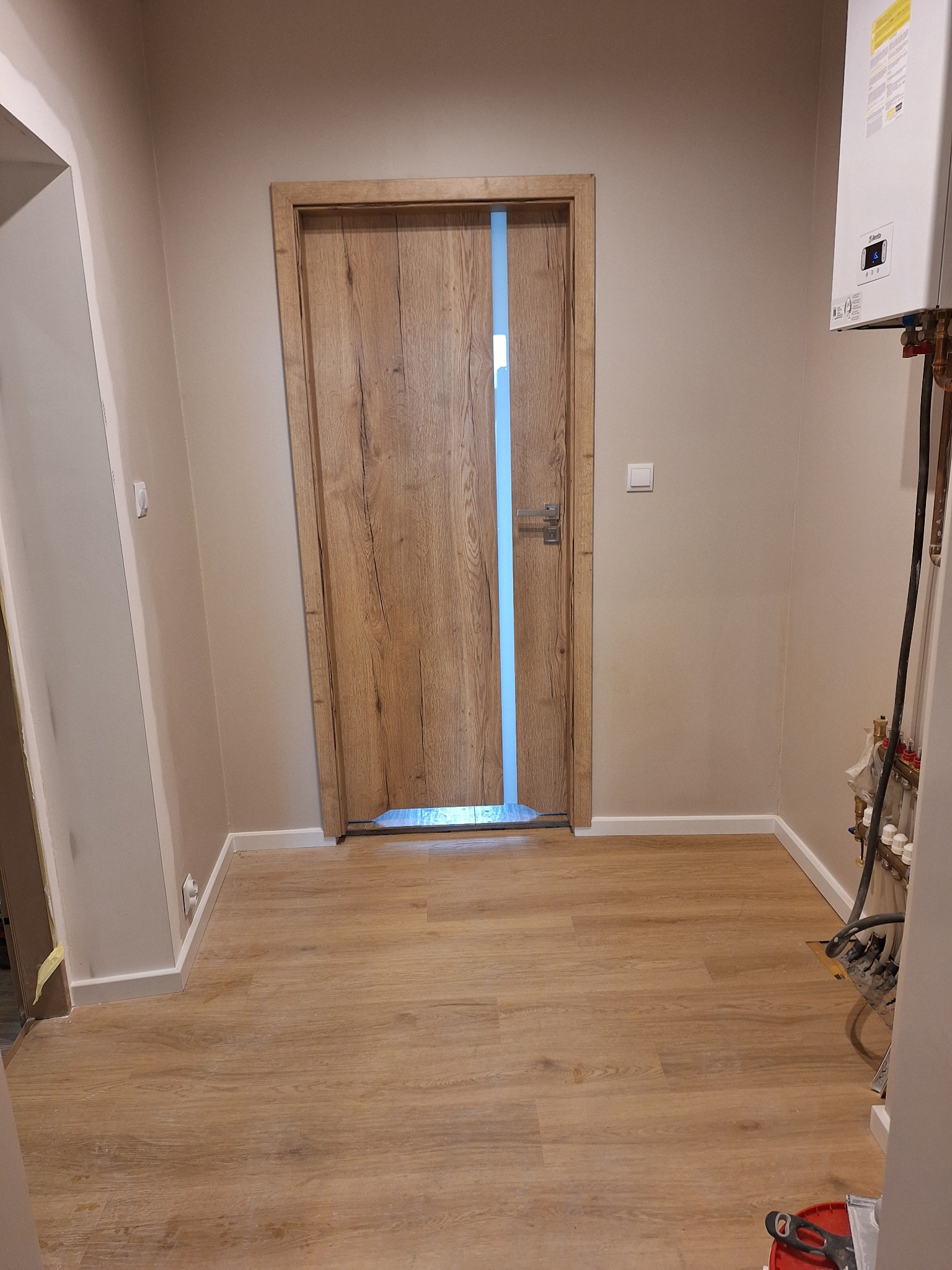 Wykończony korytarz z drewnopodobną podłogą i drzwiami z ościeżnicą. Ściany w odcieniach beżu, widoczny fragment instalacji grzewczej i listwy przypodłogowe.