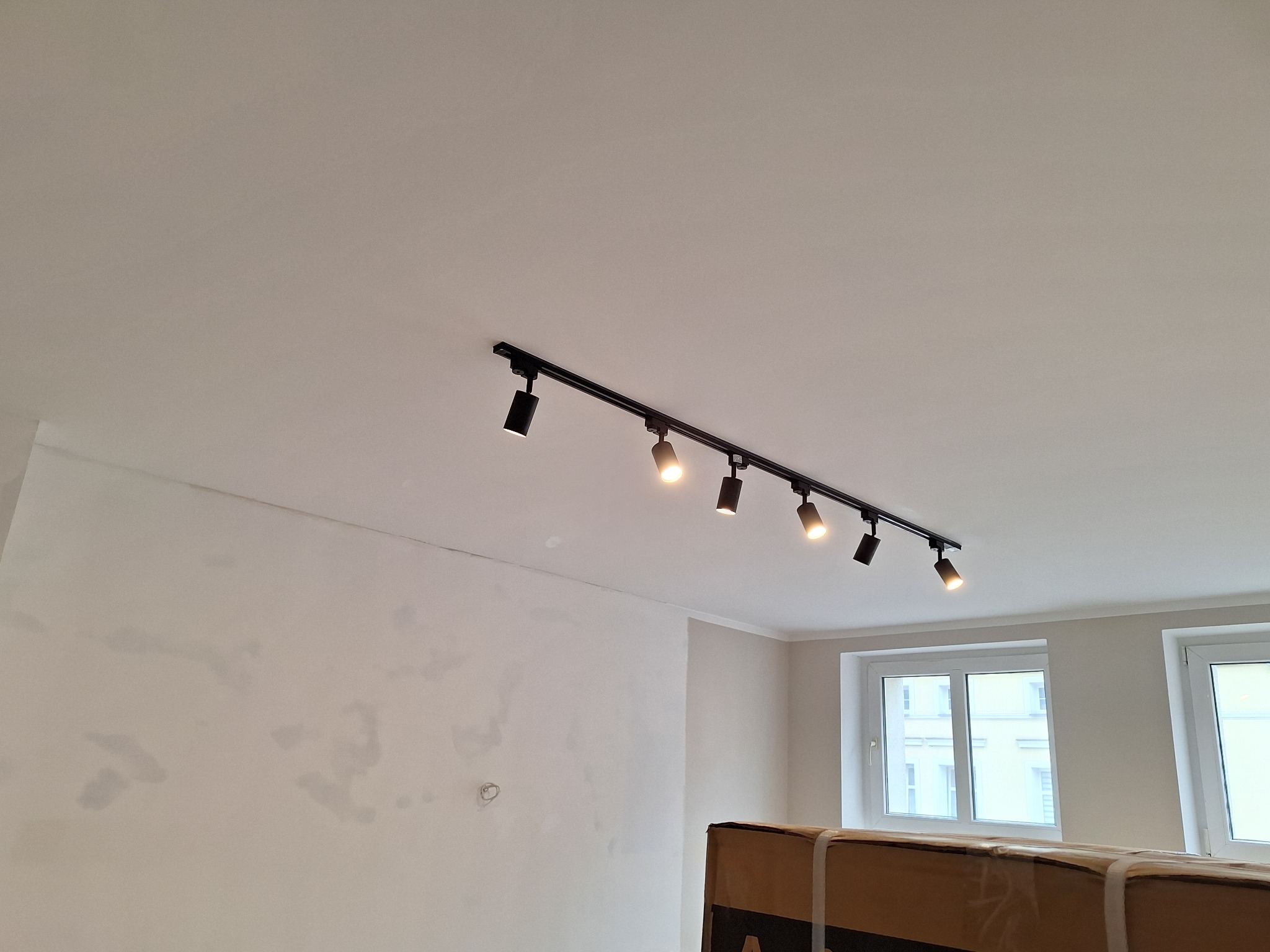 Białe wnętrze w trakcie remontu: ściany po gładzi, sufit z szynoprzewodem i reflektorami. Widoczne okna i fragment kartonu na podłodze. Realizacja oświetlenia.