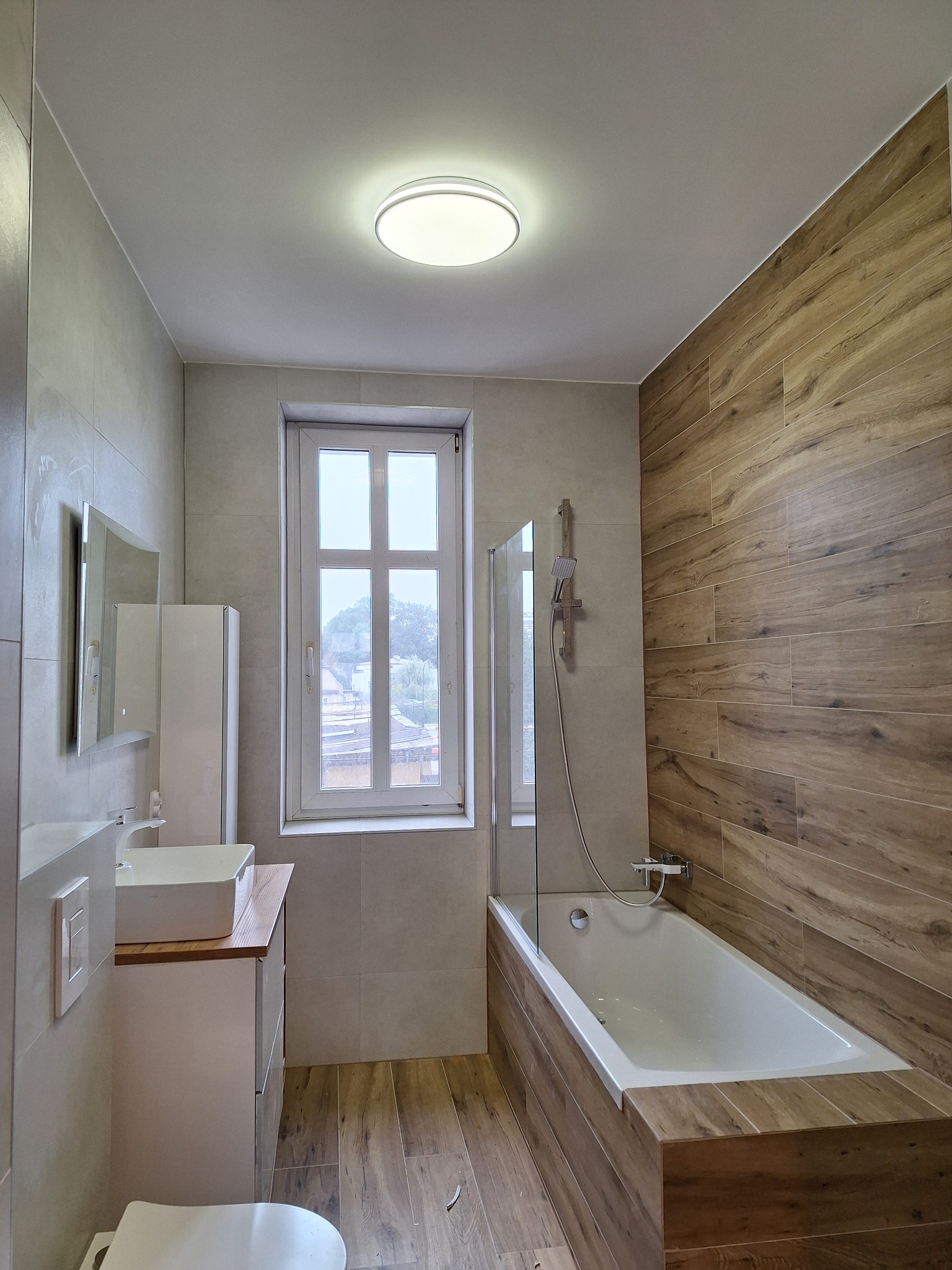 Nowoczesna łazienka z drewnopodobnymi płytkami na ścianie i podłodze, biała armatura, okno z widokiem na zewnątrz. Oświetlenie sufitowe.