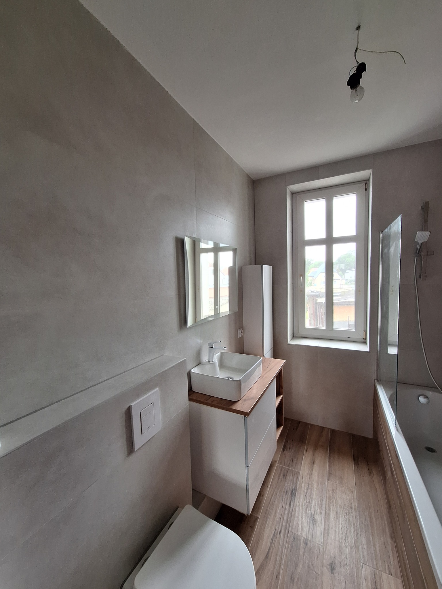 Nowoczesna łazienka po remoncie: szare płytki na ścianach, drewnopodobna podłoga, biała umywalka z szafką i lustro. Widoczne okno i wanna z parawanem.