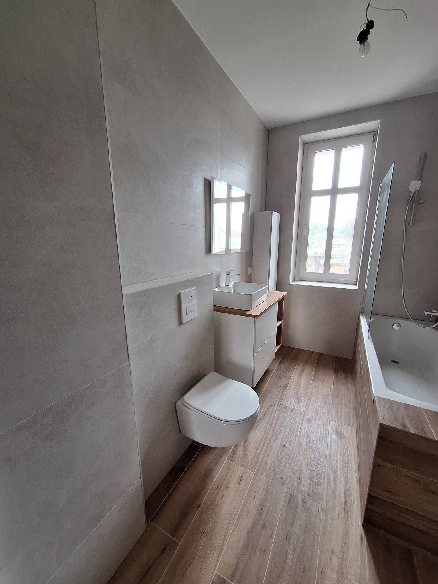 Nowoczesna łazienka po remoncie w Połczynie-Zdroju. Płytki imitujące drewno na podłodze, szare płytki na ścianach, wisząca toaleta i wanna z parawanem.