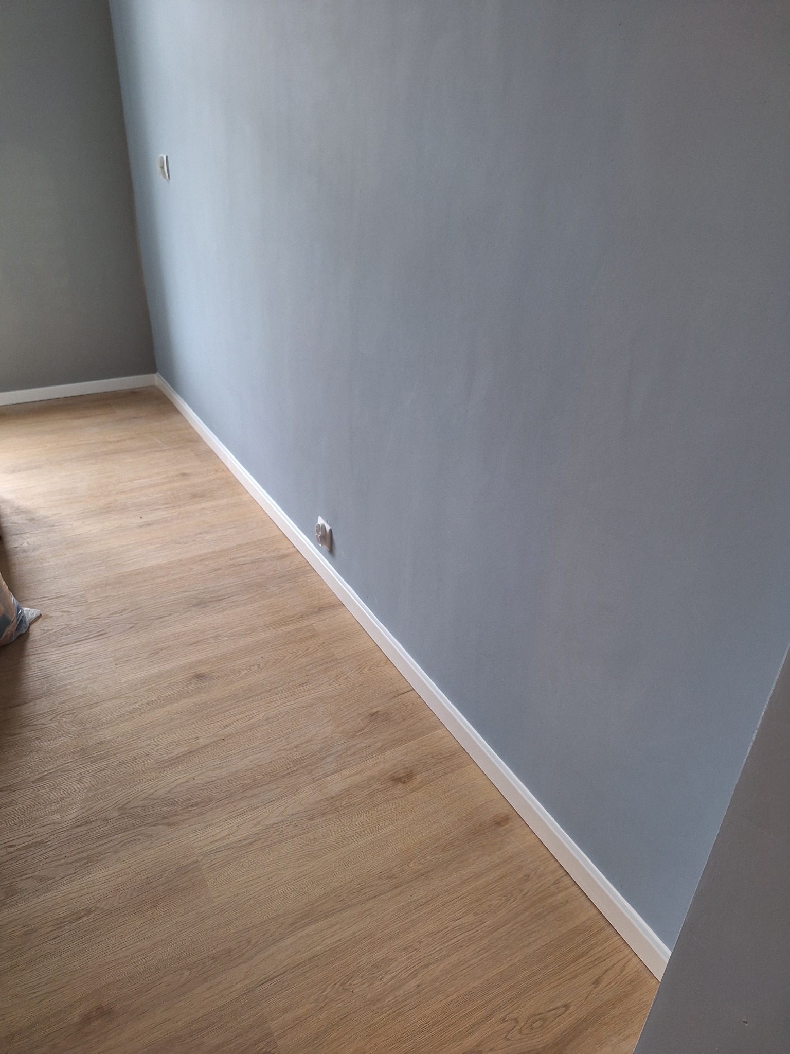 Pomalowane na szaro ściany, drewniana podłoga i białe listwy przypodłogowe w pustym pokoju z gniazdkiem elektrycznym. Nowoczesne wykończenie wnętrza.