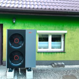 NNTT Tomasz Król - Zewnętrzna jednostka pompy ciepła JBG HT, zamontowana na betonowych podstawach przy zielonej ścianie domu z brązowymi drzwiami i oknem z roletą.