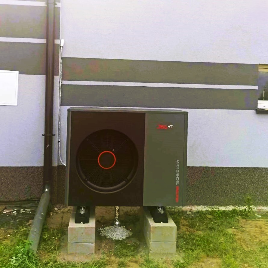 Zewnętrzna jednostka nowoczesnej, czarno-szarej instalacji grzewczej, zamontowana na betonowych bloczkach przy szarej ścianie budynku z widoczną rurą spustową.