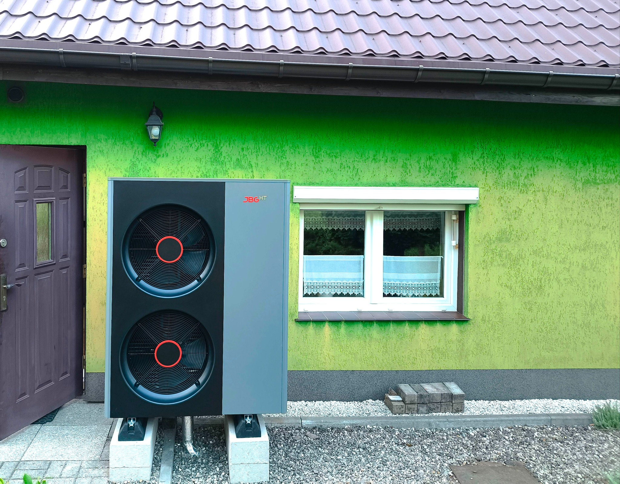 Zewnętrzna jednostka pompy ciepła JBG HT, zamontowana na betonowych podstawach przy zielonej ścianie domu z brązowymi drzwiami i oknem z roletą.