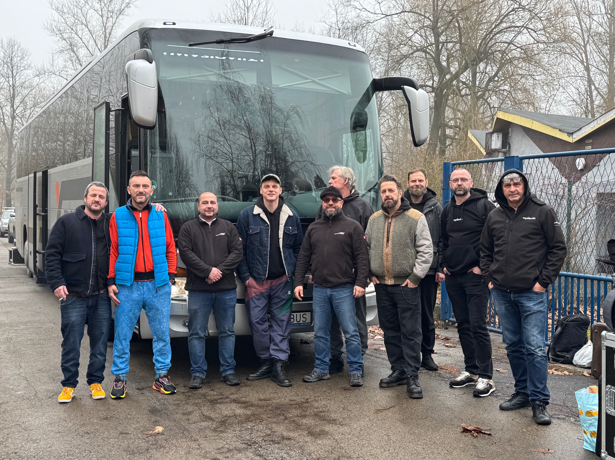 Grupa mężczyzn stoi przed srebrnym autokarem z widoczną tablicą rejestracyjną BUS, na tle szary krajobraz i budynki.