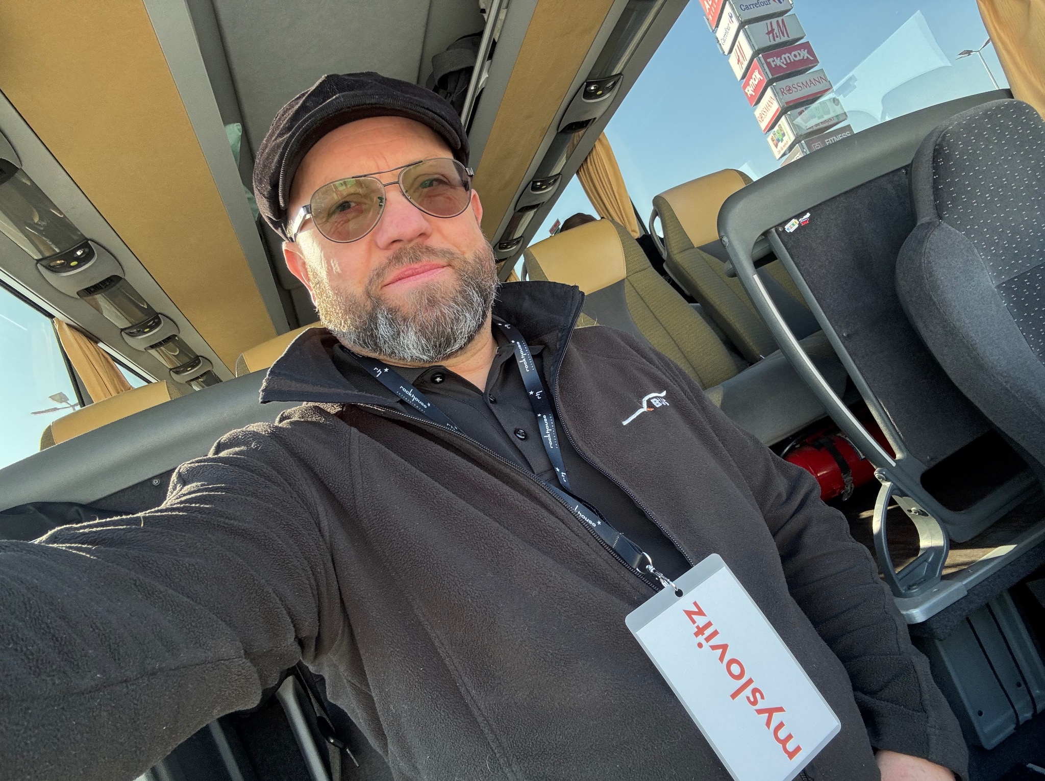 Mężczyzna w autobusie, widoczne siedzenia, za oknem znaki reklamowe sklepów, na szyi identyfikator firmy.