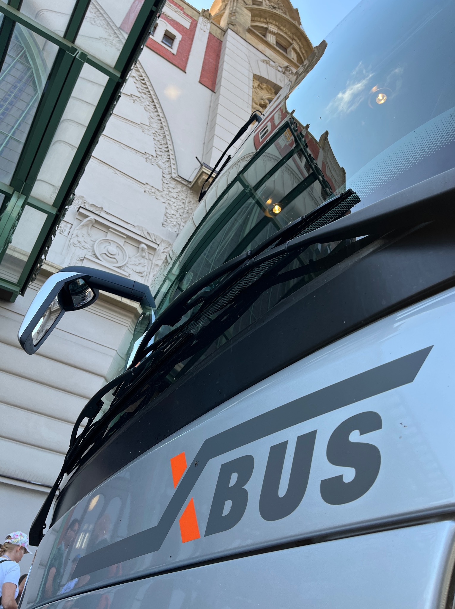 Szary autobus XBUS z pomarańczowym akcentem na tle budynku w stylu secesyjnym z elementami zdobień i przeszklonym dachem przystanku.