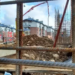 Widok przez zbrojenie na plac budowy z widoczną konstrukcją budynku w trakcie wznoszenia, stertą ziemi i pompą do betonu w tle.