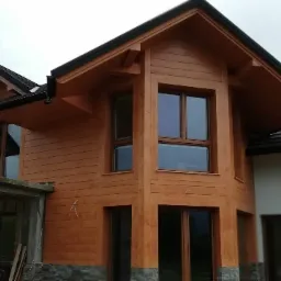 Elewacja drewniana domu w trakcie budowy, widoczne deski elewacyjne, okna z szybami, fragment dachu z rynnami i surowy betonowy element konstrukcyjny z kamienną okładziną.